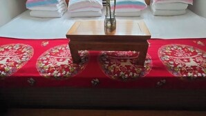 Ein ordentlich bezogenes Bett mit rotem floralem Überwurf, ein kleiner Holztisch und sauber gefaltete Handtücher.