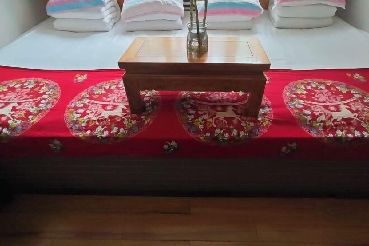 En pænt redt seng med et rødt blomstret sengetæppe, et lille træbord og pænt foldede håndklæder.