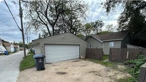 A residential garage with a gravel driveway, a wooden fence, and a trash bin.