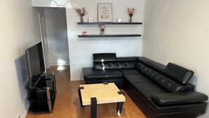 A modern living room with a black leather sectional sofa, a wooden coffee table, and a flat-screen TV on a black stand. There are shelves above the sofa with decorative items and a framed motivational quote.