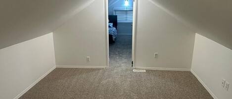 Una habitación tipo ático estrecha, con alfombra, paredes blancas y techo inclinado.
