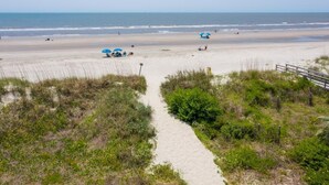 A beach with a sandy path leading to the water, some umbrellas, and a wooden fence. - Upstairs at Ocean | 4BR + Sunroom | Sleeps 8 (Isle of Palms)