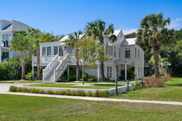 A large white house with a staircase leading to a covered porch, surrounded by palm trees and a well-maintained lawn.