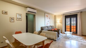 Una sala de estar con un sofá, una mesa de comedor con sillas y una mesita. Hay una ventana con cortinas, una lámpara y obras de arte en las paredes.