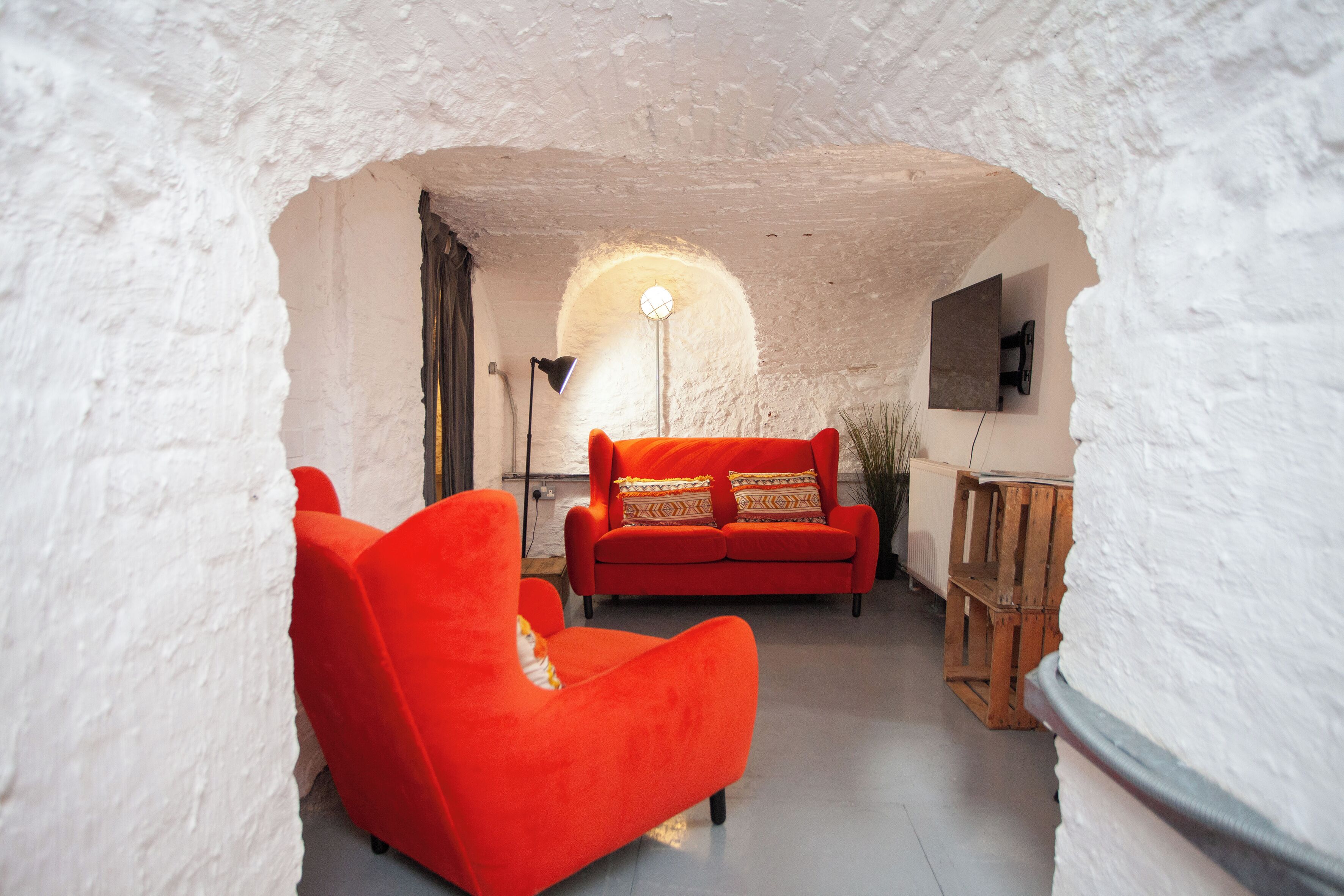 Un salon chaleureux avec un canapé rouge et une lampe, agrémenté d’un mur blanc à texture et d’une caisse en bois.