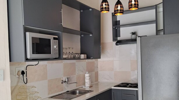 A compact kitchen with a microwave, washing machine, and a refrigerator. There are hanging pendant lights and a tiled backsplash. - Hazel Haven 2 Guest House (Pretoria)