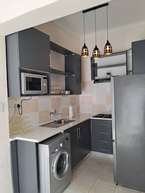 A compact kitchen with a microwave, washing machine, and a refrigerator. There are hanging pendant lights and a tiled backsplash. - Hazel Haven 2 Guest House (Pretoria)