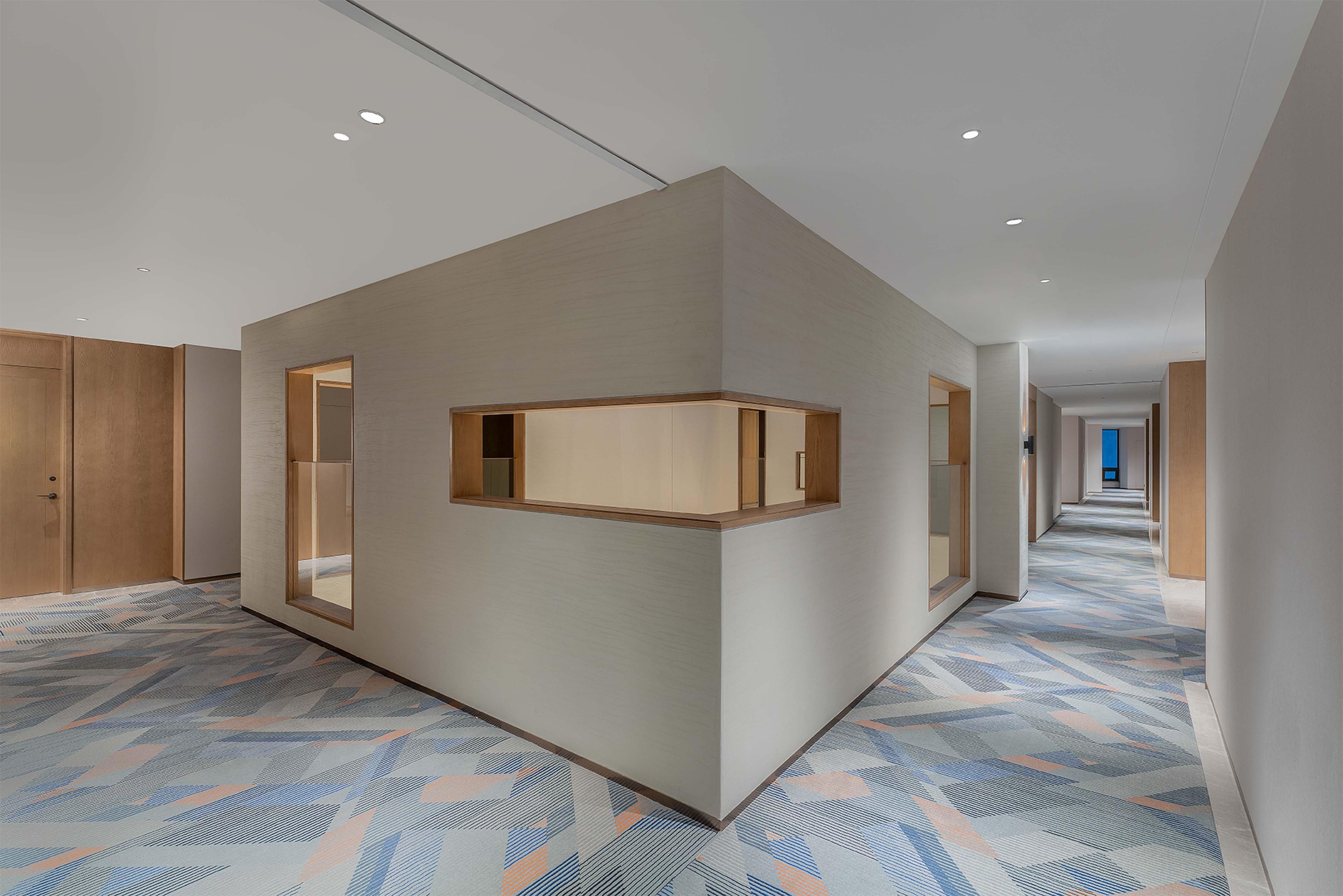 A modern hotel corridor with a geometric patterned floor, wooden paneling on the walls, and recessed ceiling lights.