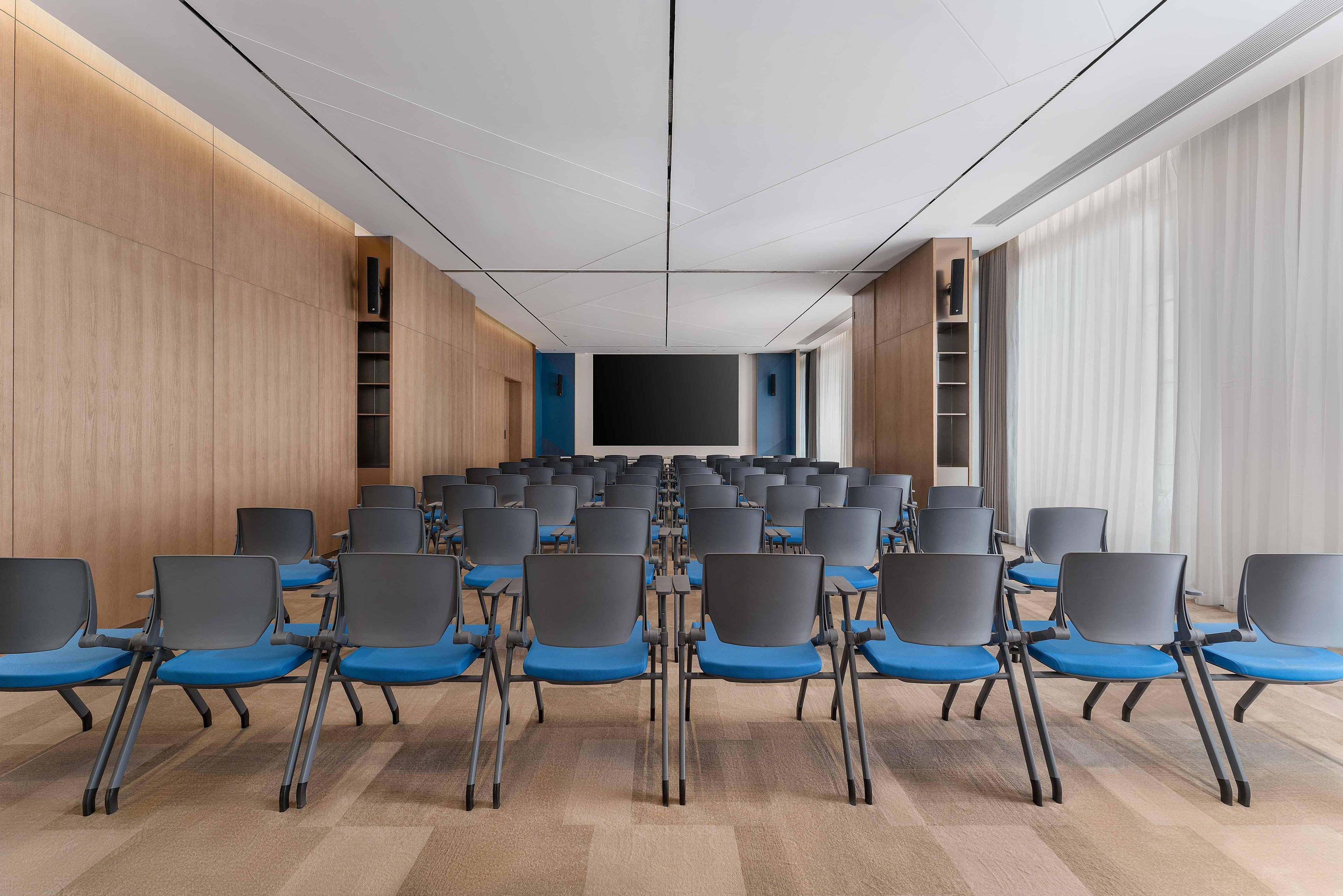 A conference room with rows of chairs, a large screen, and wooden paneling on the walls.
