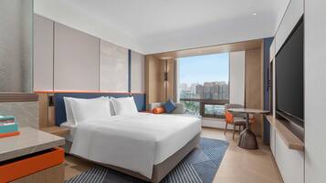 Habitación de hotel moderna con una cama grande, un escritorio, una silla, un televisor y una ventana con vistas a la ciudad.