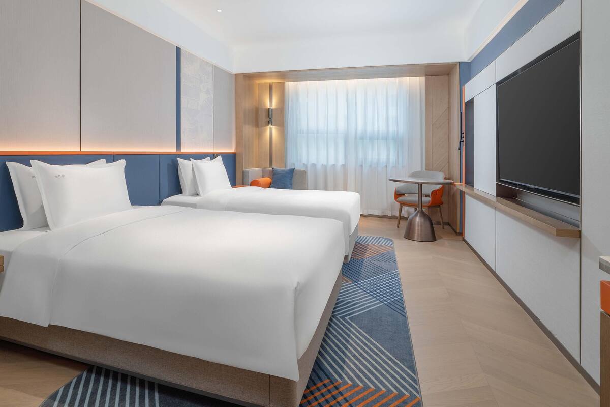Moderni hotellihuone, jossa on kaksi sänkyä, työpöytä, tuoli, televisio ja ikkuna, jossa on verhot.
