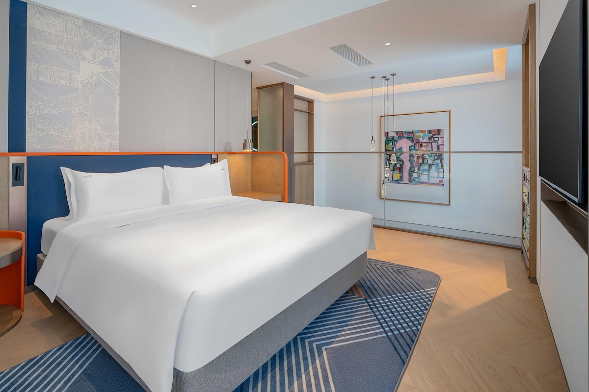 A modern hotel room with a large bed, a small seating area, a TV, and a painting on the wall.
