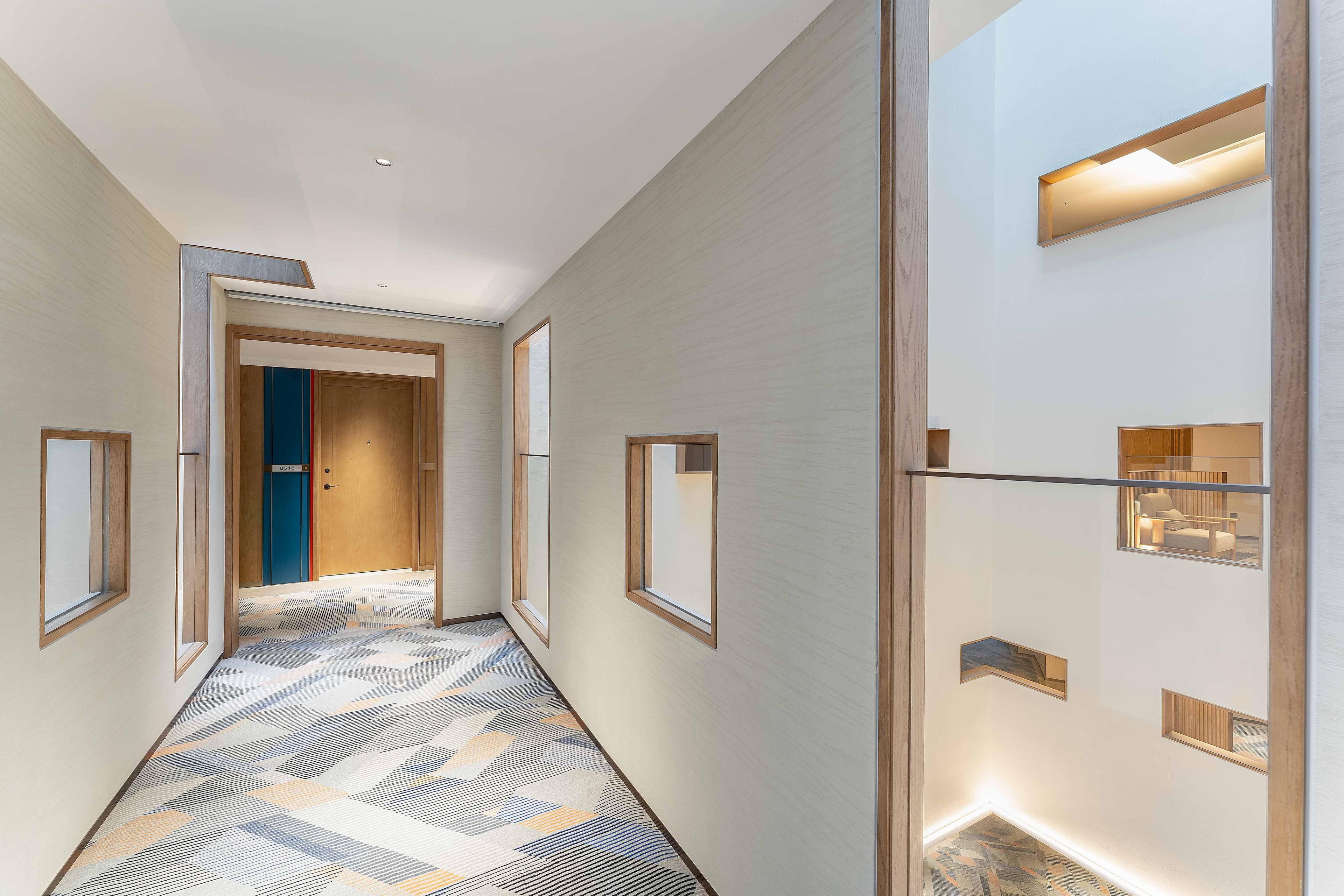 Un pasillo moderno de hotel con suelo estampado, ventanas de madera y luces empotradas.