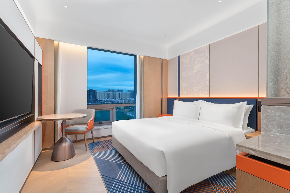 A modern hotel room with a large bed, a desk, a chair, and a TV.