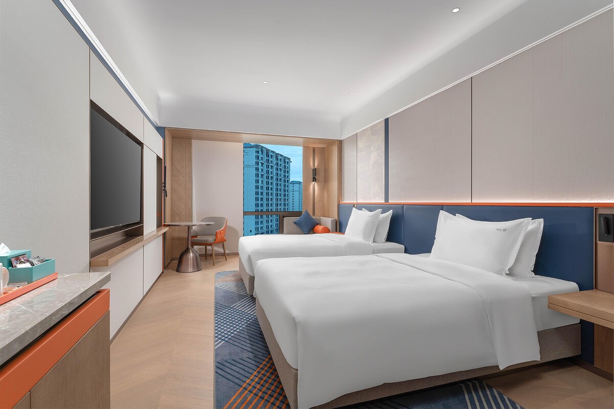 A hotel room with two beds, a TV, a desk, and a view of a cityscape.