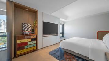 A modern hotel room with a large bed, a colorful dresser, a flat-screen TV, and a window with a view of buildings.