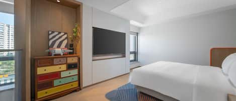 A modern hotel room with a large bed, a colorful dresser, a flat-screen TV, and a window with a view of buildings.