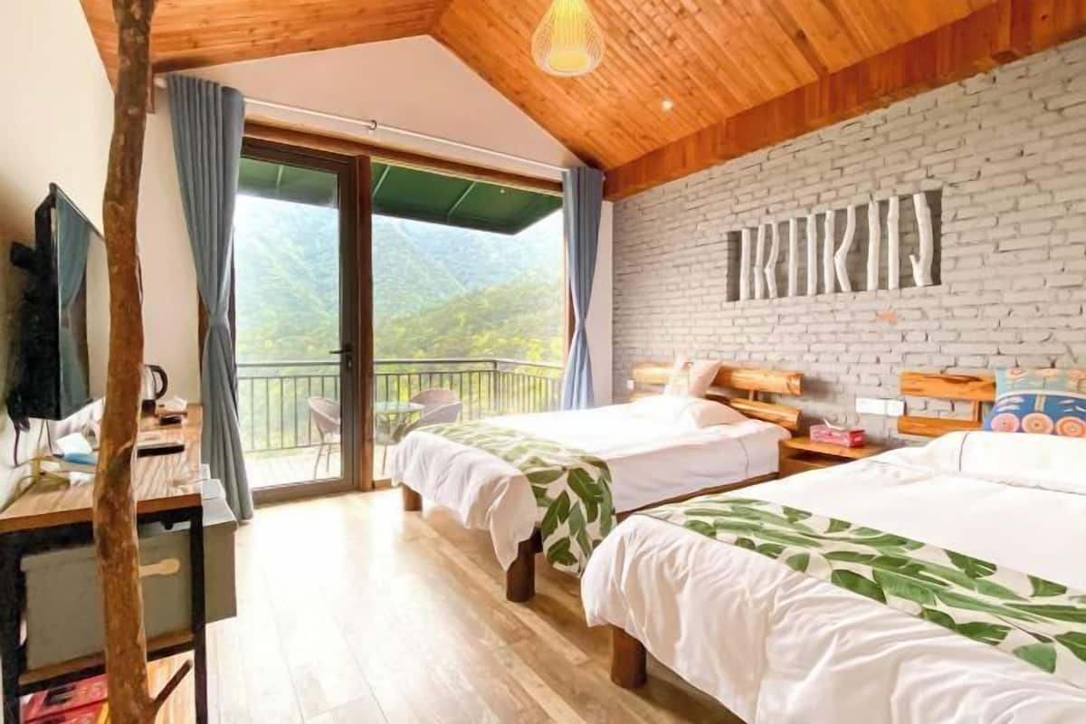 A room with two beds, a wooden desk, and a balcony with a view of the mountains.