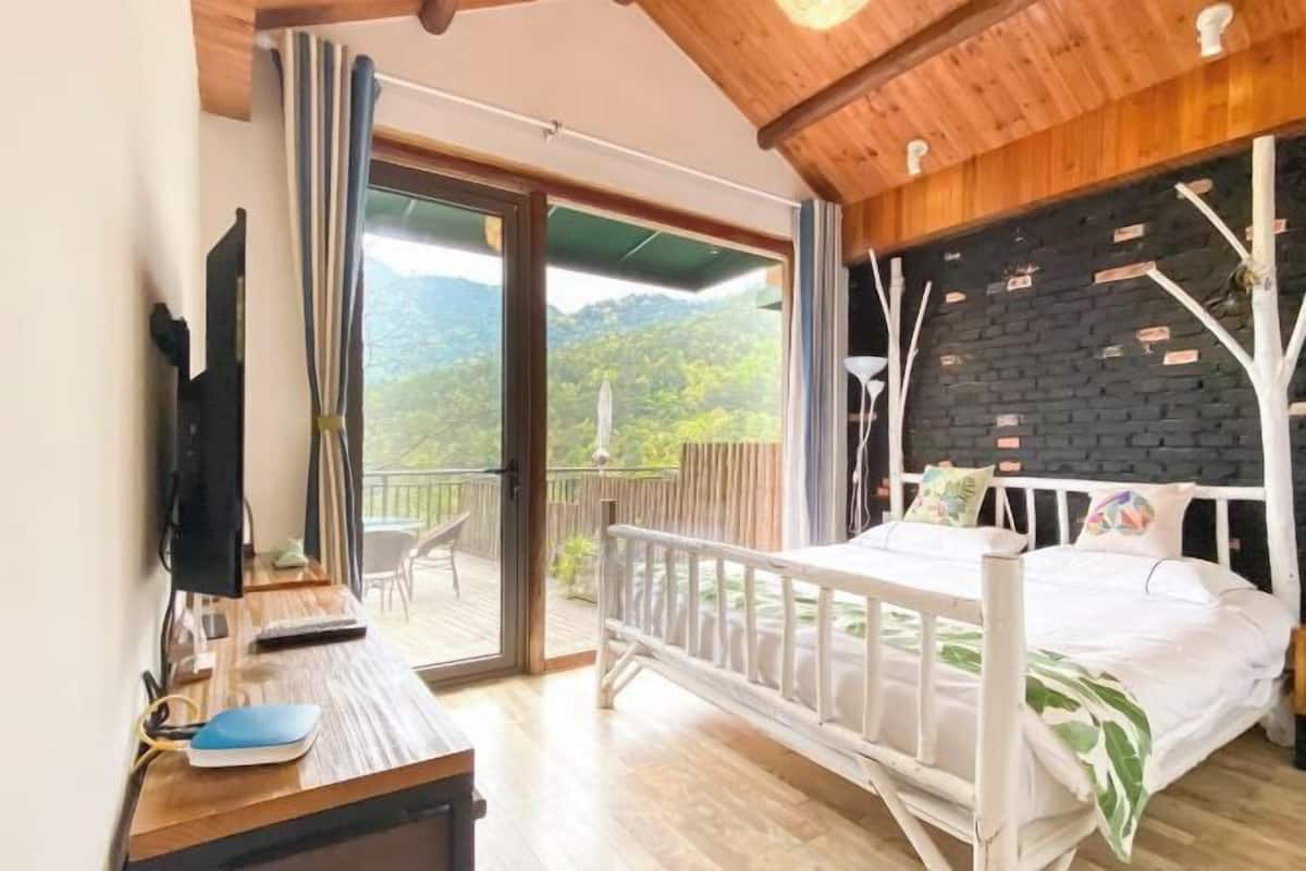 A bedroom with a bed, a desk with a computer, a TV, and a balcony with a view of the mountains.