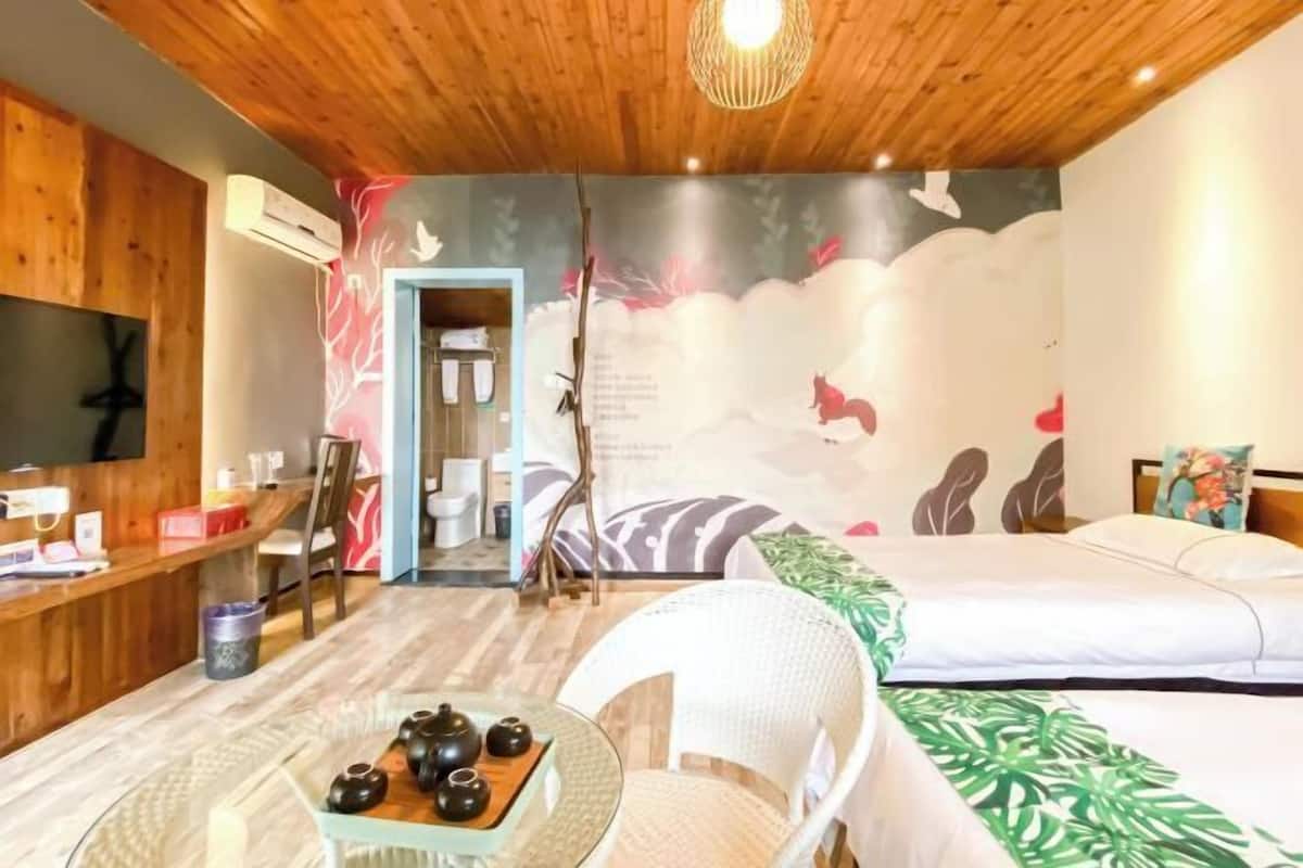 A room with a wooden ceiling, a bed with a green leaf-patterned bedspread, a white chair, a small table with teapots, and a wall with a nature-themed mural.