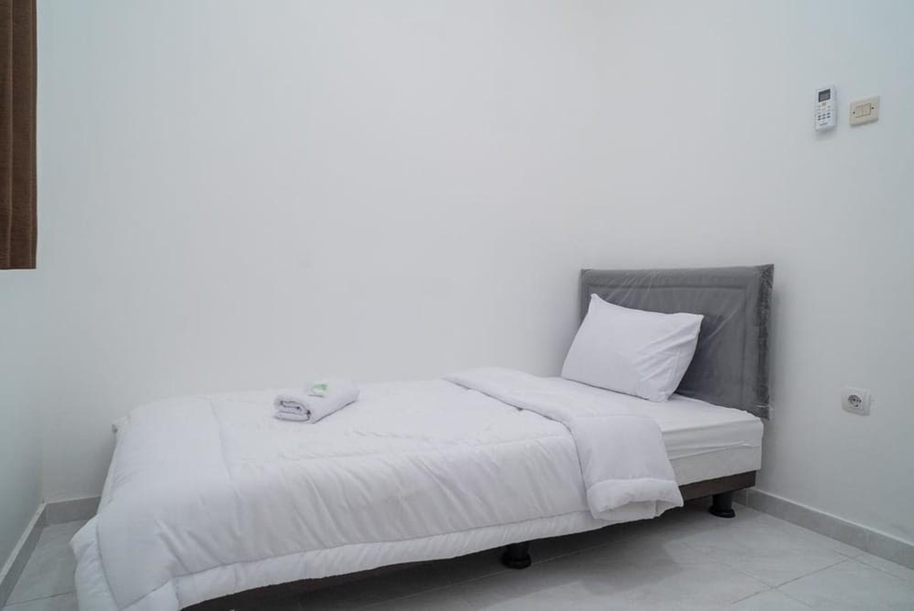 A small, neatly made bed with white bedding and a grey headboard in a minimalistic room with white walls and a tiled floor.
