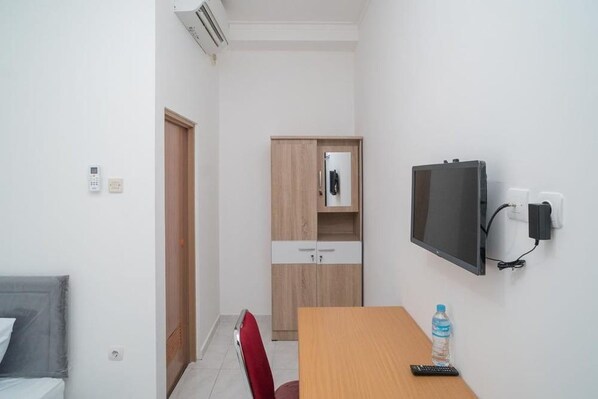 Una habitación pequeña y bien iluminada con una cama, una mesa de madera con un control remoto, una botella de agua, un televisor montado en la pared y un armario con espejo.