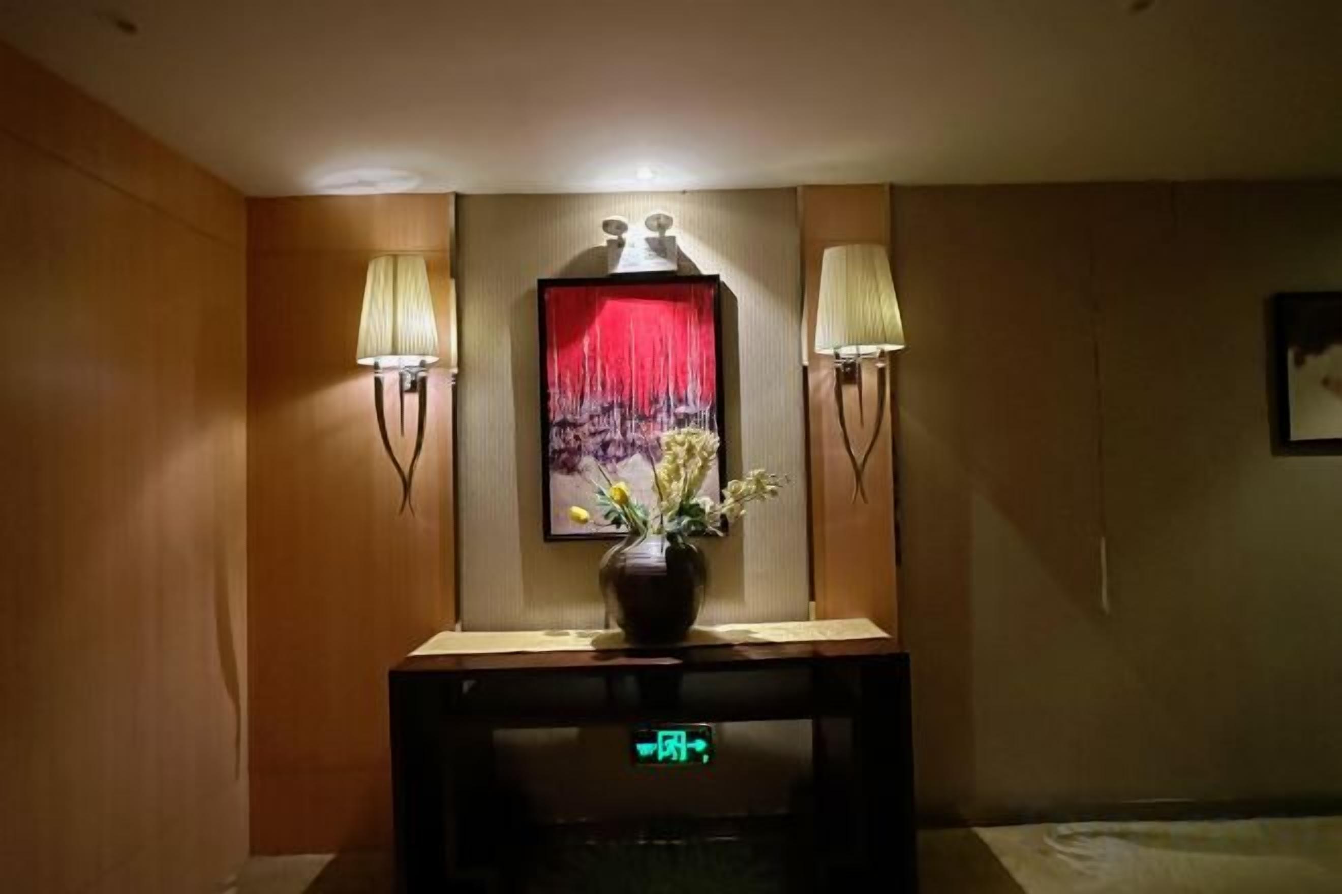 A hotel reception area with a reception desk, two wall-mounted lamps, a vase with flowers, and a red curtain.