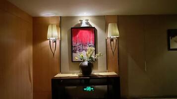 A hotel reception area with a reception desk, two wall-mounted lamps, a vase with flowers, and a red curtain.