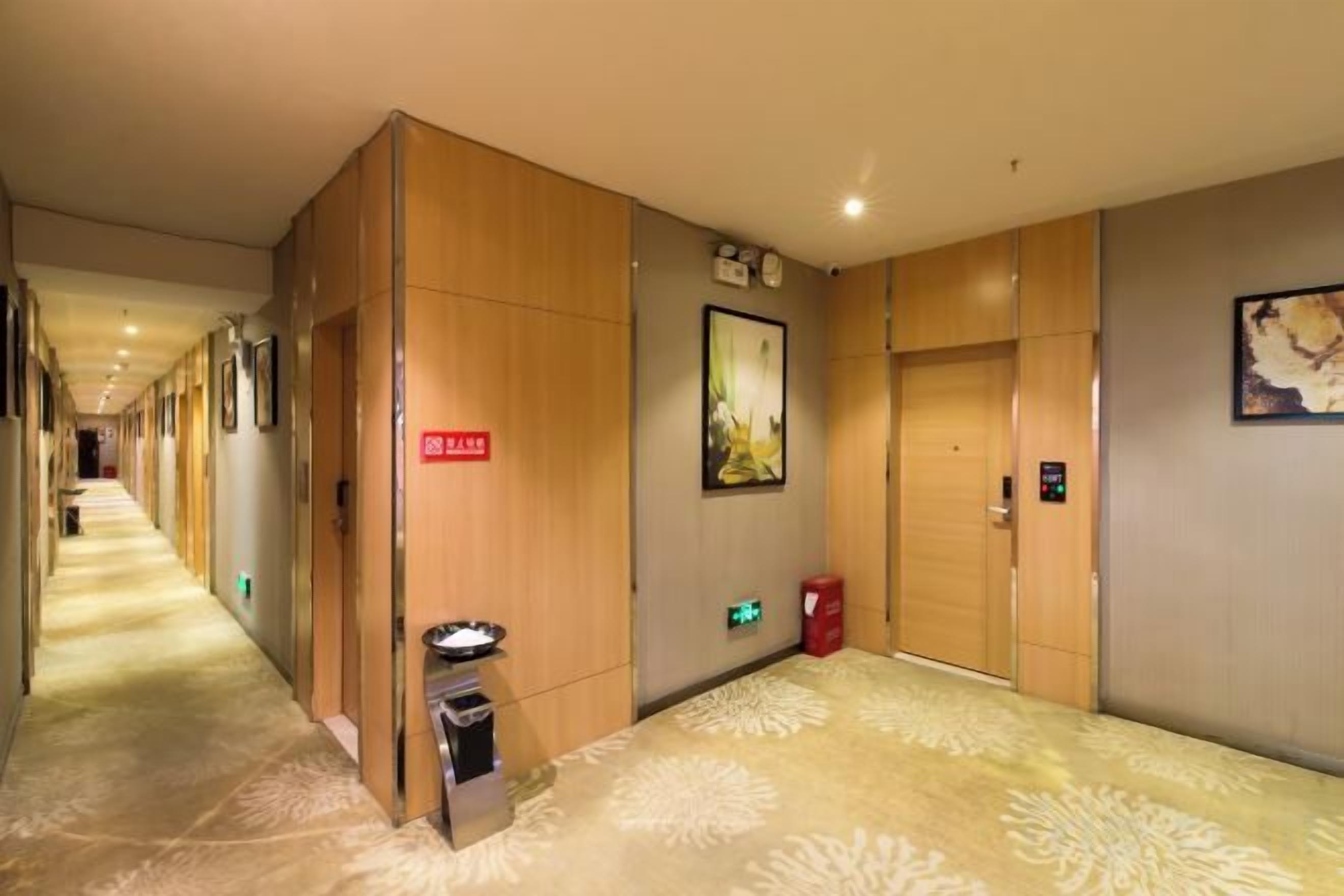 A hotel corridor with wooden paneling, a trash bin, a red trash bin, a green exit sign, and a door with a keypad lock.