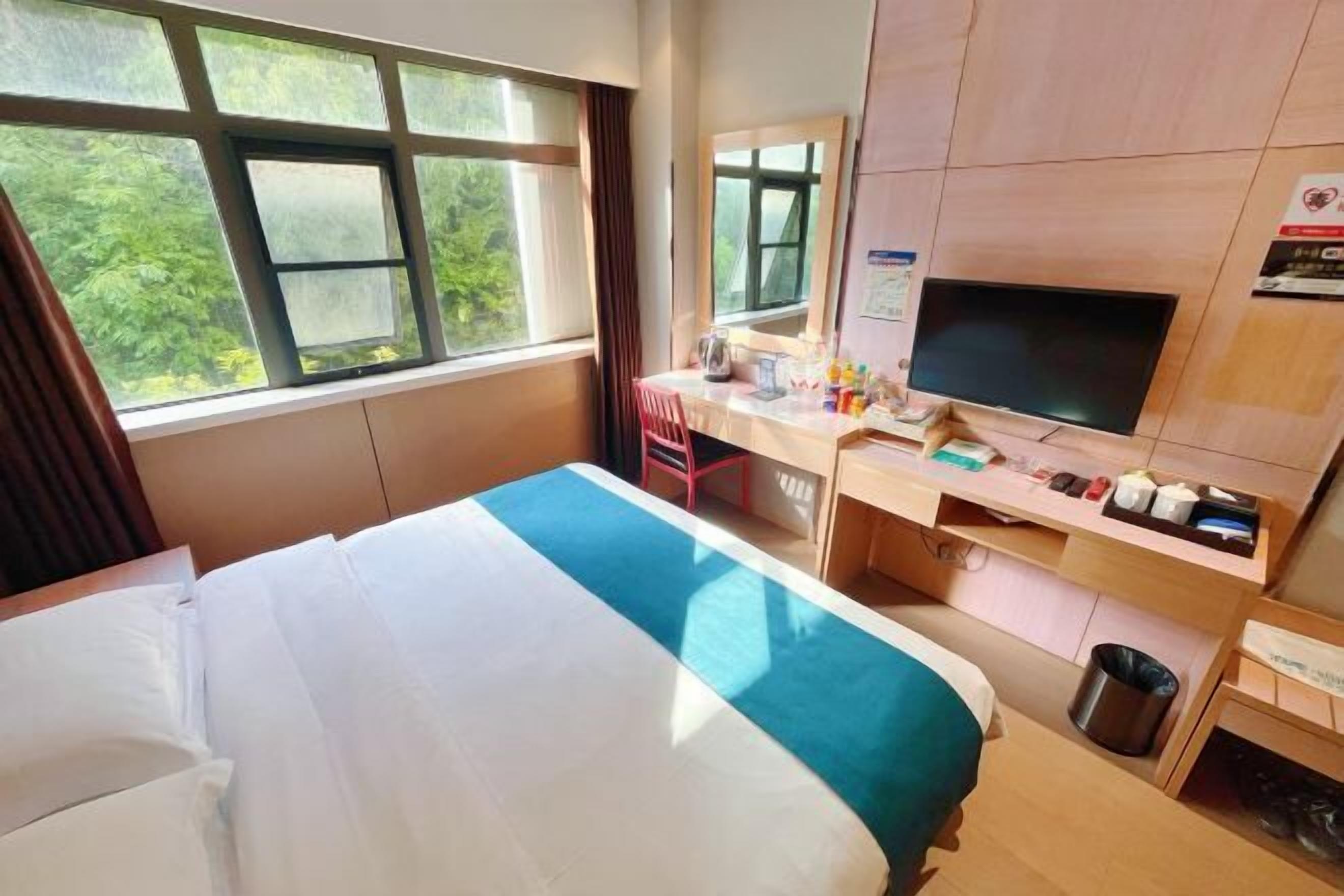 Ein Hotelzimmer mit Bett, Schreibtisch, Fernseher und einem Fenster mit Blick auf Grünflächen.