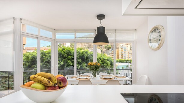 Una cocina moderna con un amplio ventanal, un cuenco de fruta sobre la encimera y una zona de comedor con una mesa dispuesta para dos personas.
