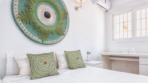 Un dormitorio con una cama, dos almohadas decorativas, una obra de arte en la pared, una lámpara de noche, una ventana y un aparador blanco.