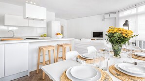 Una cocina y comedor moderna, con decoración blanca, un ventanal grande, un televisor de pantalla plana y una mesa de comedor preparada para cuatro.
