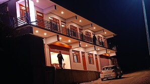 Ein zweigeschossiges Haus mit Balkon, ein Auto vor der Fassade und eine Person auf dem Balkon.