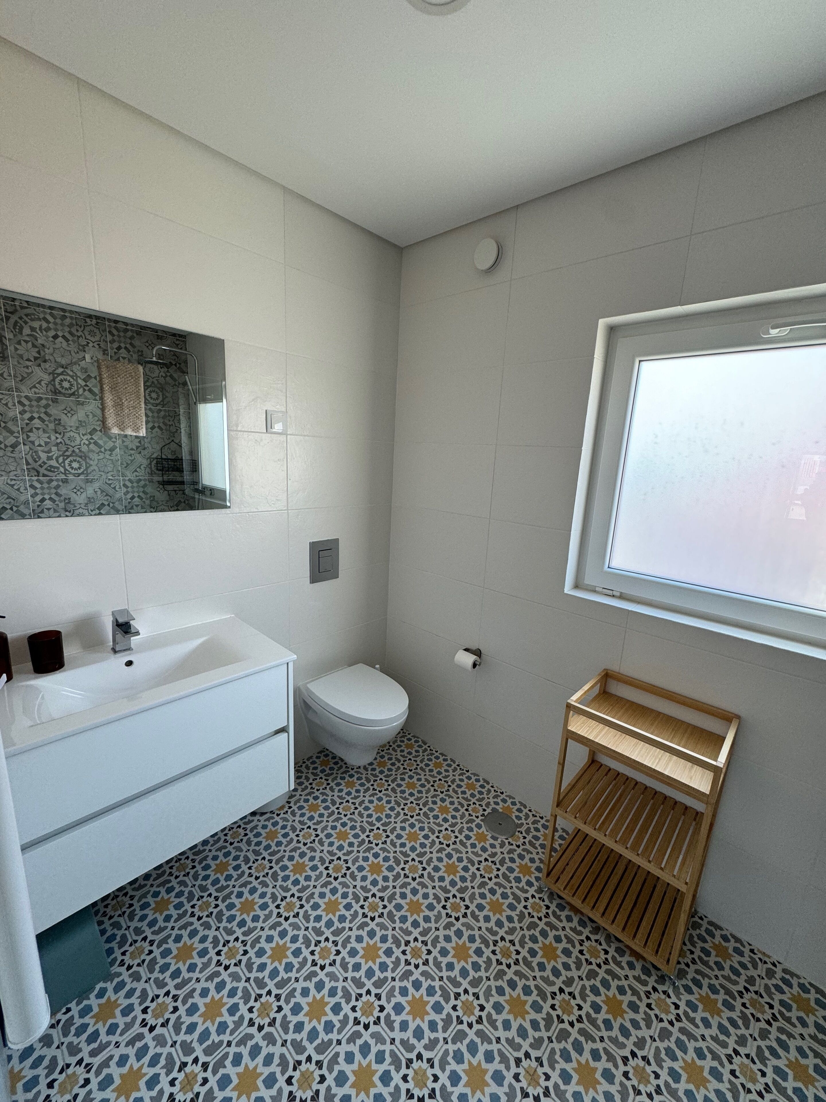 Une salle de bain avec un lavabo blanc, des toilettes et un miroir au-dessus du lavabo. Il y a une fenêtre sur le mur et un carrelage décoratif au sol.