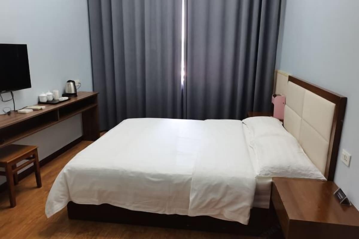 A hotel room with a bed, a desk with a television, a kettle, and a bedside table with a pink item.