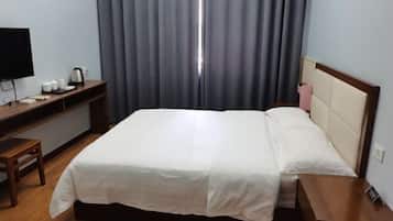 A hotel room with a bed, a desk with a television, a kettle, and a bedside table with a pink item.