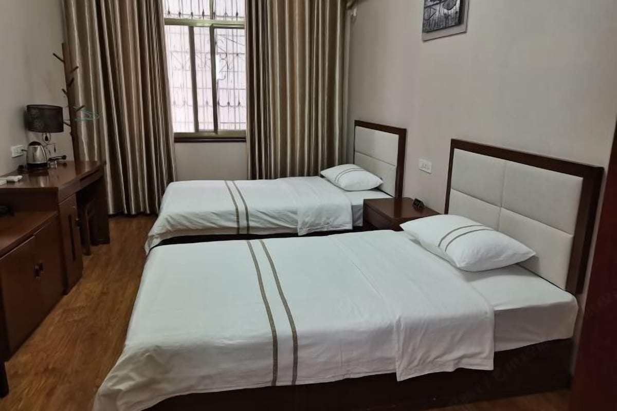 A hotel room with two single beds, a desk with a lamp, a window with curtains, and a wall-mounted picture.