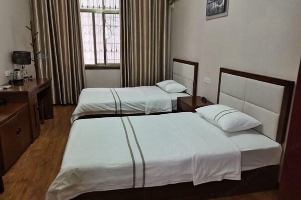 A hotel room with two single beds, a desk with a lamp, a window with curtains, and a wall-mounted picture.