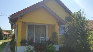 A yellow house with a red roof, a front porch with two white chairs, and a garden with red flowers. - Apartment Čeliković (Draz)