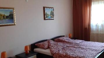 A bedroom with a bed, two bedside tables, a nightstand, a window with curtains, and two framed pictures on the wall.