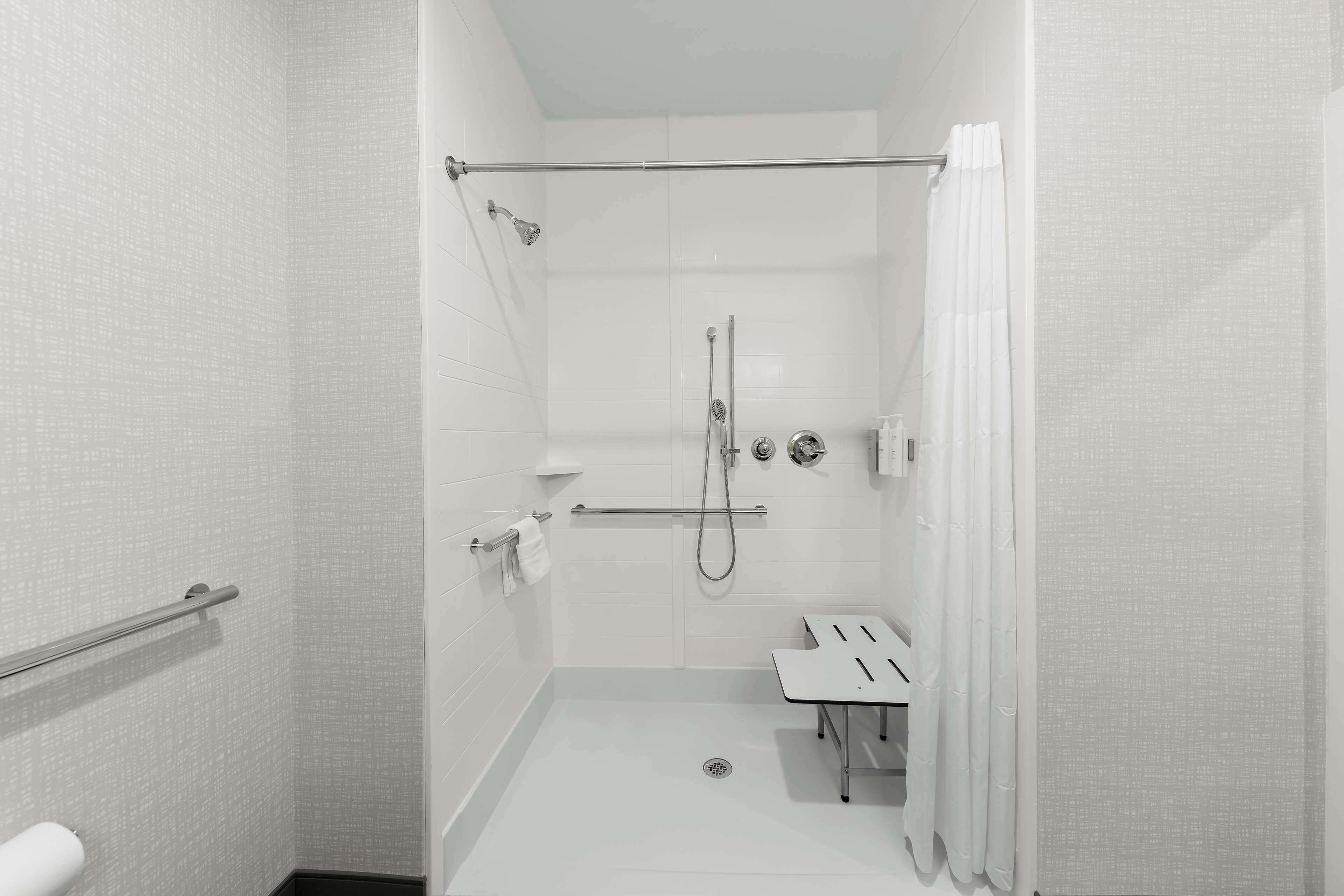 A modern, accessible shower area with a glass door, a handrail, a showerhead, and a handheld shower attachment.