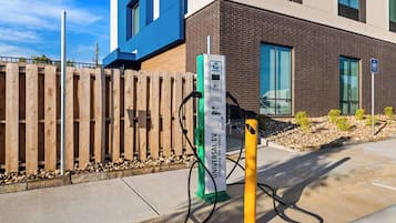 Estação de carregamento para veículos elétricos, com cerca de madeira e um edifício moderno ao fundo.