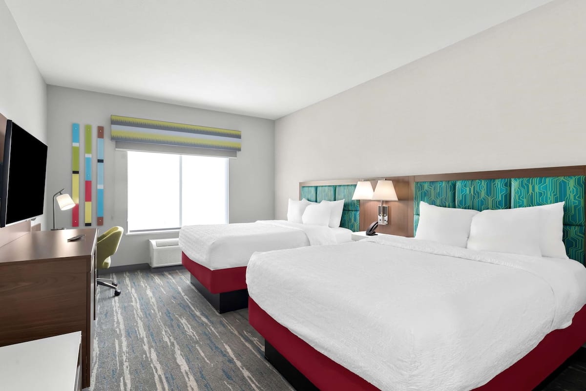Habitación de hotel con dos camas, un escritorio, una silla, un televisor y una ventana con persianas.