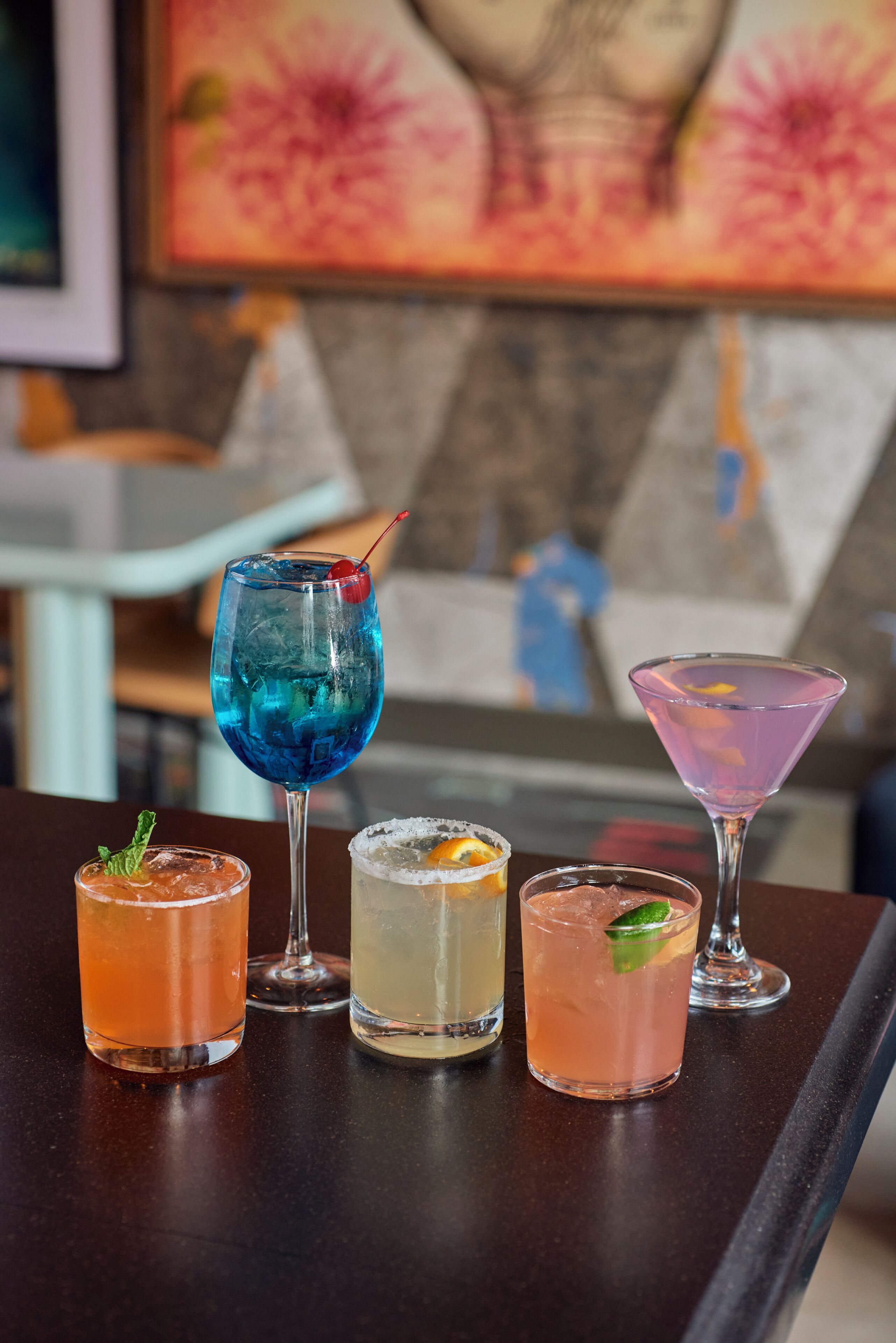 Eine Bar mit fünf verschiedenen Cocktails, einem Gemälde im Hintergrund und einem weißen Stuhl.