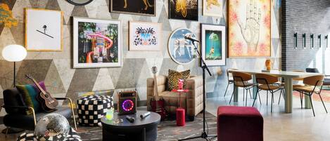 A modern living room with a geometric wall design, a variety of framed artworks, a guitar, a disco ball, and a red ottoman.