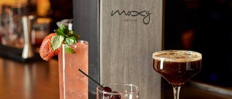 A bar counter with three different drinks, a sign with the word 'mozy' on it, and a blurred background suggesting a hotel setting.