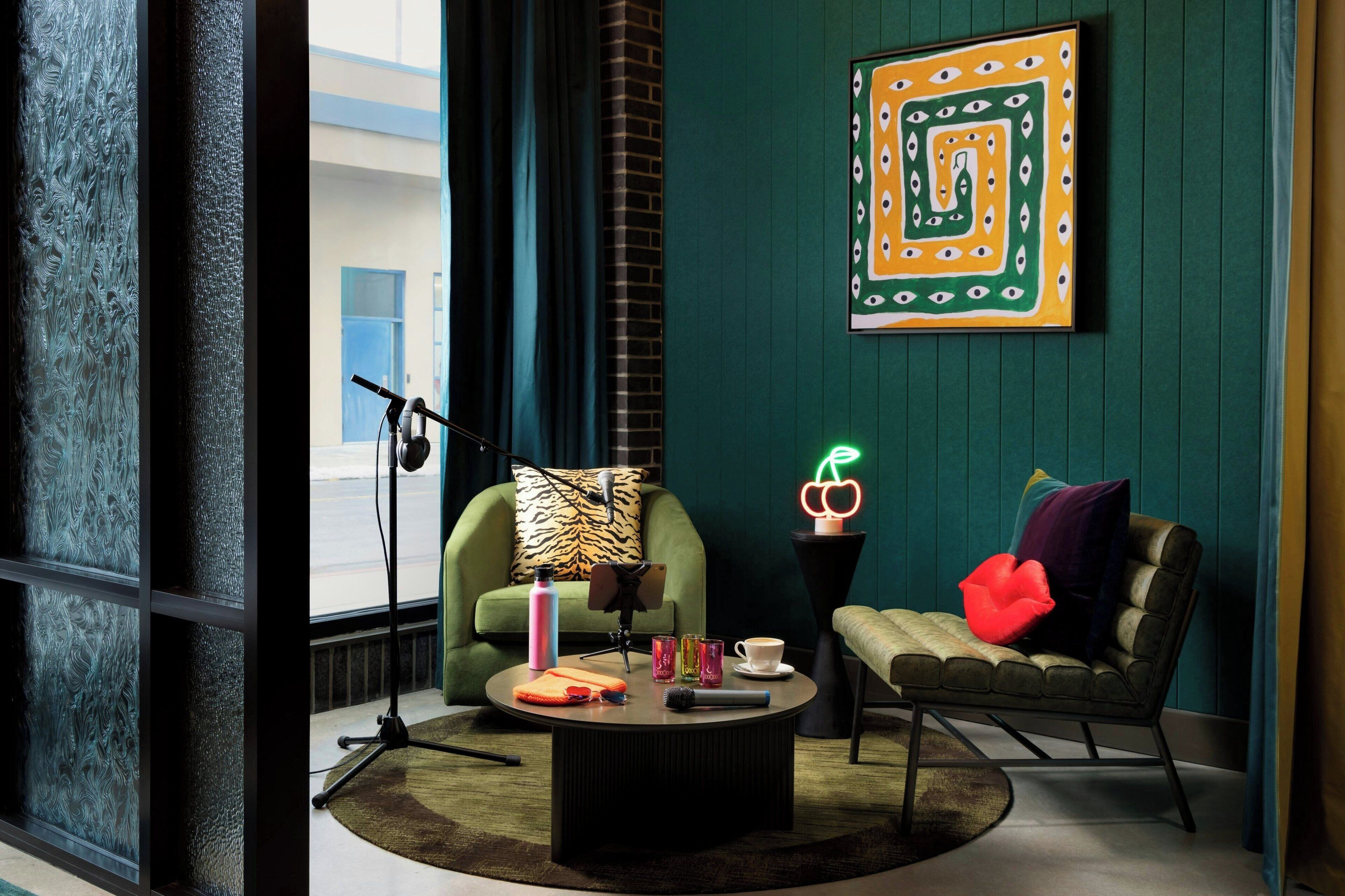 A cozy room with a green wall, a framed abstract art piece, a green armchair, a round coffee table with various items, and a window with frosted glass.