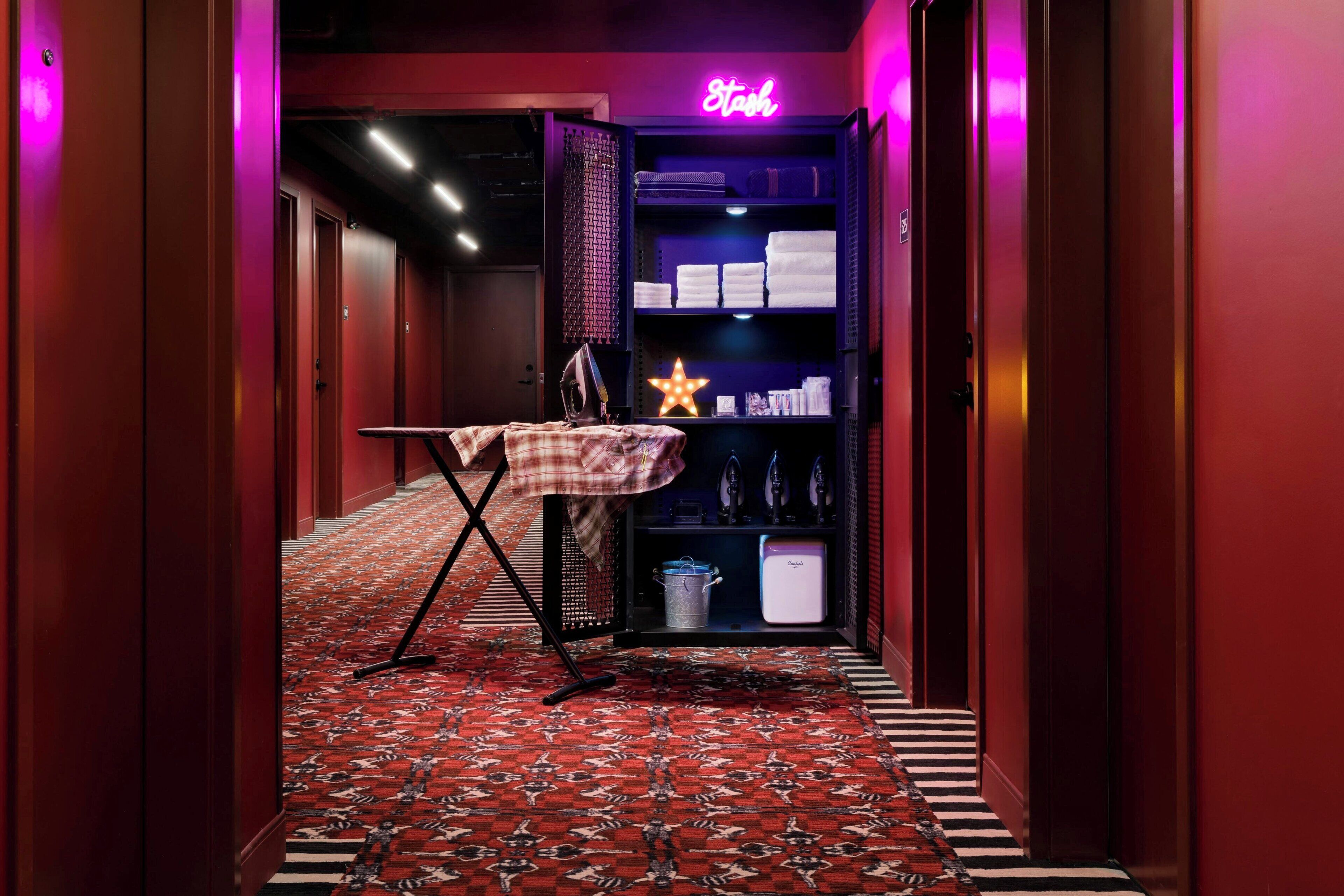 A hotel hallway with a red carpet, a neon sign reading "Stash," and a shelf with towels and toiletries.
