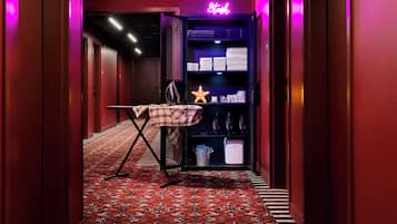 A hotel hallway with a red carpet, a neon sign reading "Stash," and a shelf with towels and toiletries.